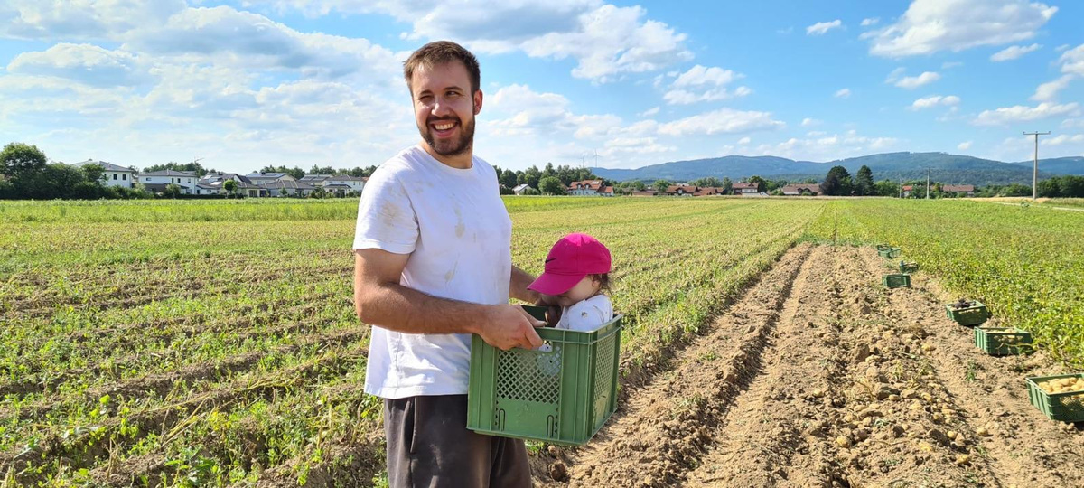 KI generiert: Ein Mann steht lächelnd auf einem Feld und trägt ein Kind in einer grünen Kiste.