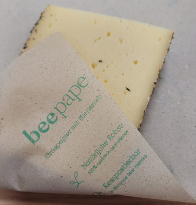 KI generiert: Ein Käse in umweltfreundlichem Bienenwachspapier verpackt. Text: "beepape, Gropapier mit Bienenwxc..."