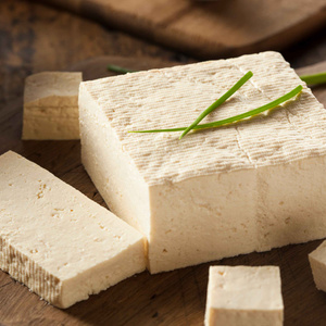 KI generiert: Das Bild zeigt einen Block Tofu, der auf einem Holzbrett liegt, mit einigen geschnittenen Stücken und dekoriert mit Schnittlauch. Der Tofu hat eine cremige Farbe und eine feste Textur.