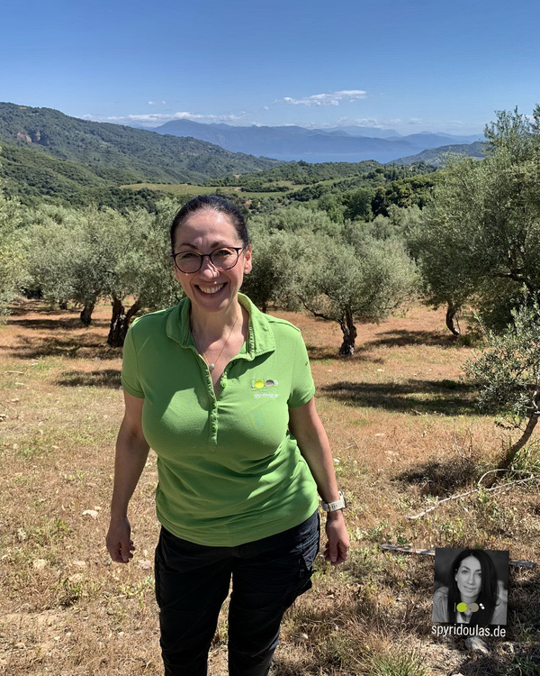 KI generiert: Eine Frau steht lächelnd in einem Olivenhain mit einer Berglandschaft im Hintergrund. Text: spyridoulas.de