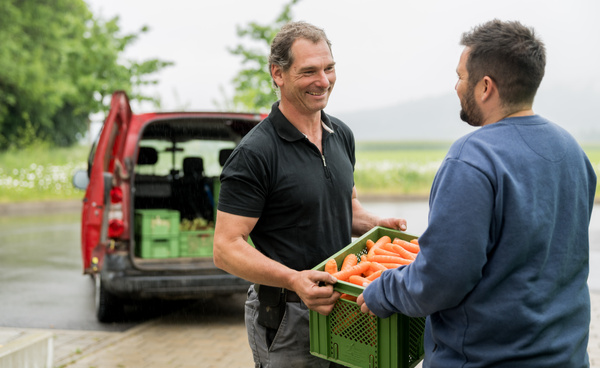 KI generiert: Zwei Männer tauschen draußen eine Kiste mit frischen Karotten aus. Im Hintergrund steht ein roter Lieferwagen.