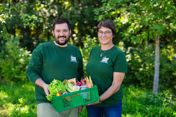 KI generiert: Zwei Personen halten eine Kiste mit frischem Obst und Gemüse in einem grünen Umfeld.