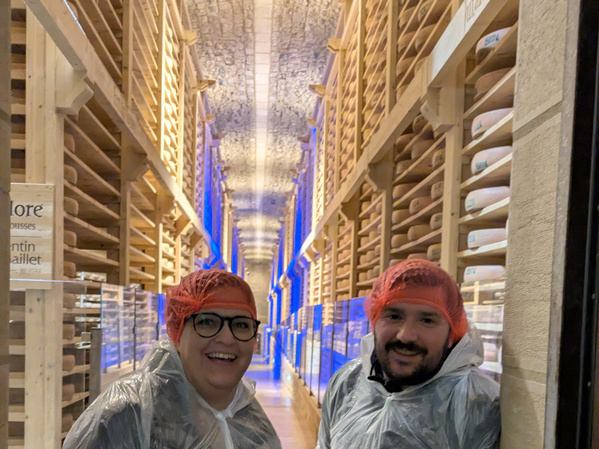 KI generiert: Zwei Personen in Schutzkleidung in einem Käsekeller mit Regalen voller Käseleibe.