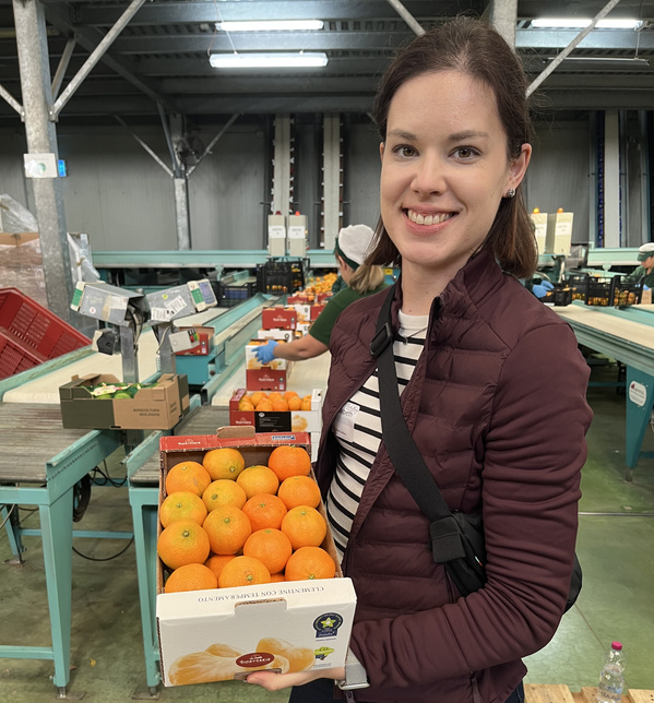 KI generiert: Eine Frau hält eine Kiste mit Orangen in einer Lagerhalle.