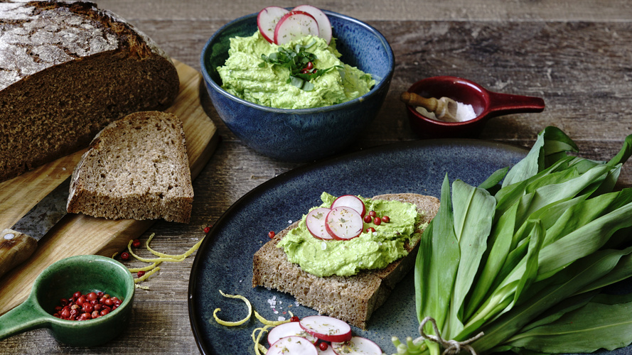 KI generiert: Ein Brot mit Bärlauchaufstrich, Radieschen und Gewürzen, daneben Bärlauchblätter und ein Laib Brot.