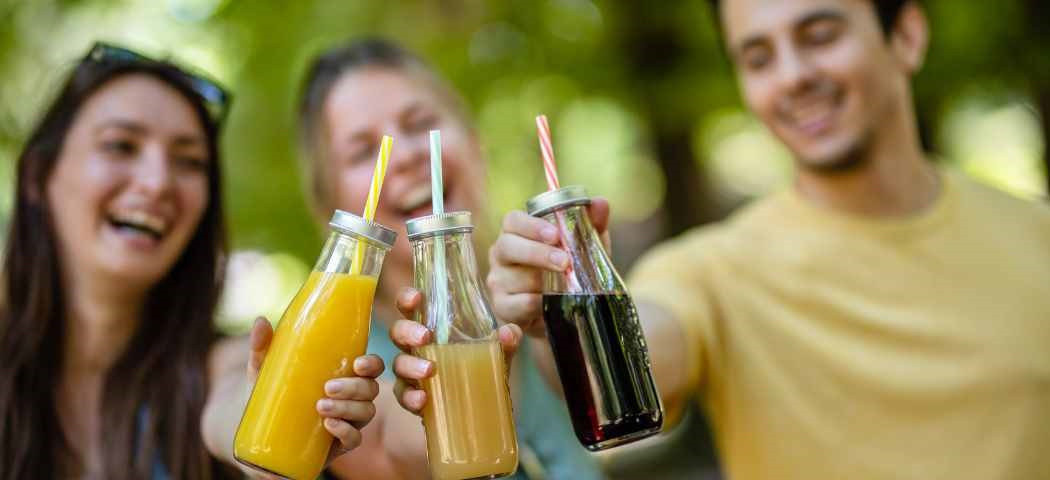 KI generiert: Drei Menschen stoßen draußen mit Flaschen voller farbiger Getränke an.