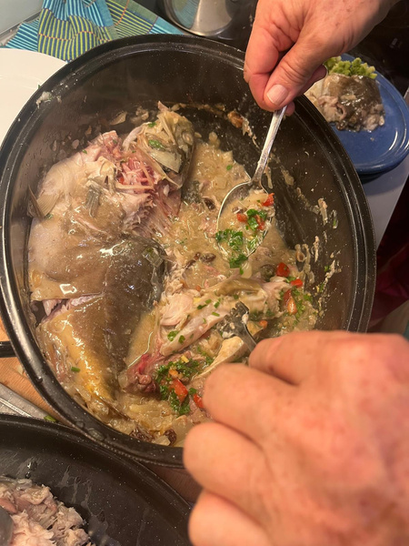 KI generiert: Das Bild zeigt eine Person, die mit einem Löffel in einem Topf kocht, in dem Fische und Gemüse in Flüssigkeit zubereitet werden. Der Hauptinhalt ist somit ein Kochvorgang mit frischem Fisch und Zutaten.