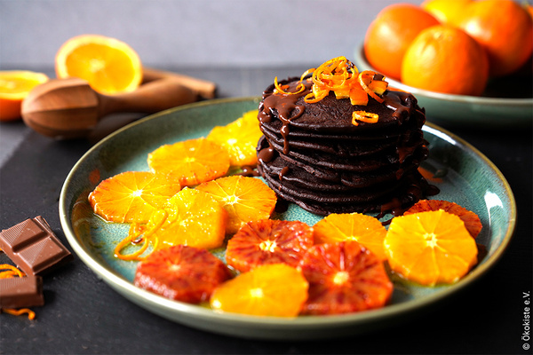 KI generiert: Das Bild zeigt einen Teller mit Schokoladenpfannkuchen, die mit Orangenscheiben und Schokoladensauce garniert sind. Im Hintergrund sind frische Orangen und ein Entsafter zu sehen.