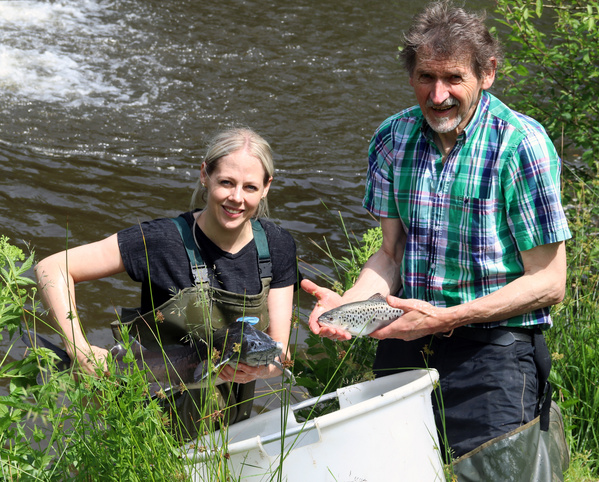 KI generiert: Zwei Personen halten Fische am Flussufer über einem weißen Behälter.