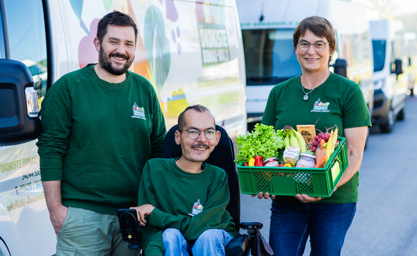 KI generiert: Drei Personen bei Lieferwagen, eine hält eine Kiste mit frischem Gemüse. Text: "Ökokiste Domäne Fredeburg".