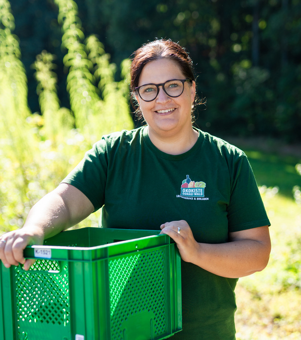 KI generiert: Person trägt grüne Kiste im Freien. Text auf Shirt: "Ökokiste Donau-Wald Lieferung & Bioladen".