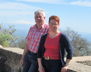 KI generiert: Zwei Personen stehen an einer gemauerten Brüstung und lächeln, während im Hintergrund eine weite Landschaft mit einem Berg sichtbar ist. Das Bild scheint an einem sonnigen Tag aufgenommen worden zu sein.