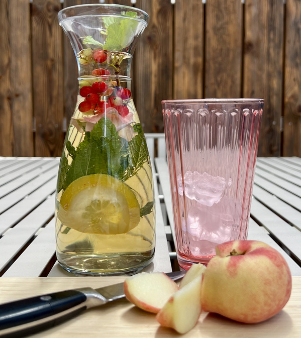 KI generiert: Eine Karaffe mit frischem Wasser, Zitrone, Minze, Beeren und einem Glas mit Eiswürfeln. Davor ein geschnittener Apfel.