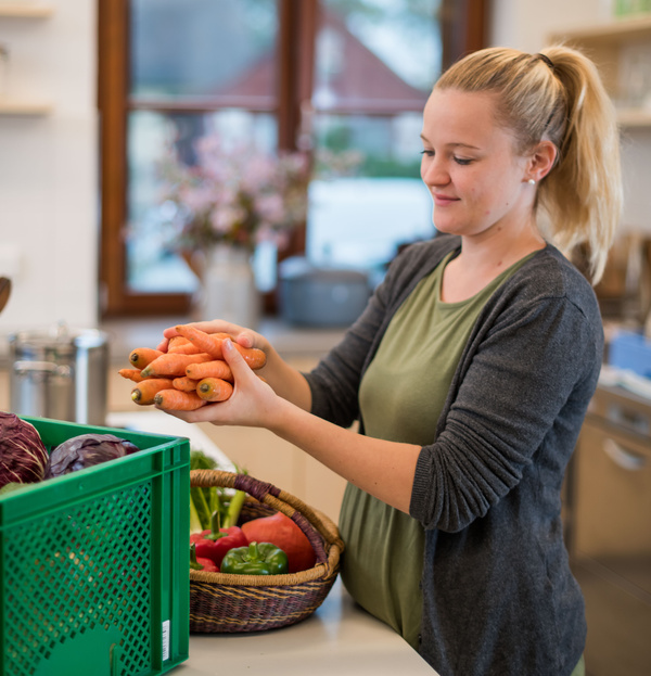 KI generiert: Eine Person hält einen Bund Karotten in der Hand und steht in einer Küche, in der auch andere frische Gemüse sichtbar sind. Der Fokus liegt auf der Zubereitung oder dem Umgang mit frischen Zutaten.
