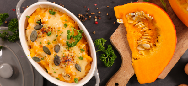 KI generiert: Das Bild zeigt ein gebackenes Kürbisgericht in einer Auflaufform, garniert mit Kernen und Kräutern. Daneben liegt ein frisches Stück Kürbis auf einem Schneidebrett.