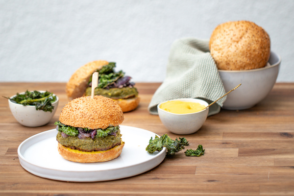 KI generiert: Das Bild zeigt einen veganen Burger mit grünen Zutaten und Brötchen mit Sesam, serviert auf einem Holzuntergrund. Im Hintergrund sind weitere Burgerzutaten, ein kleiner Napf mit Dip und eine Schüssel mit Brötchen zu sehen.