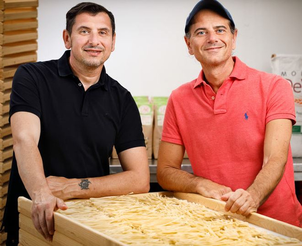 KI generiert: Zwei Männer stehen lächelnd hinter einem Holzgestell mit frischer Pasta.