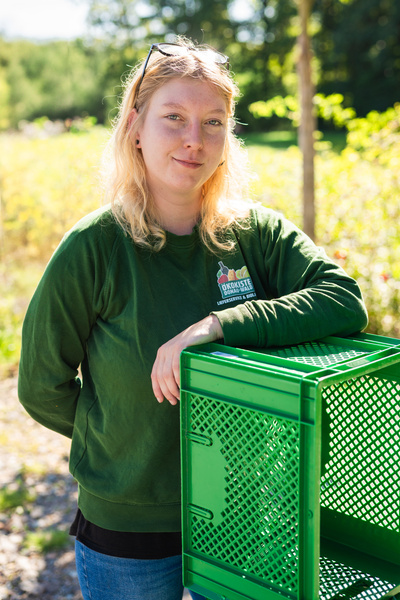 KI generiert: Person in grünem Oberteil lehnt an einem grünen Korb im Freien. Text: "Ökokiste".