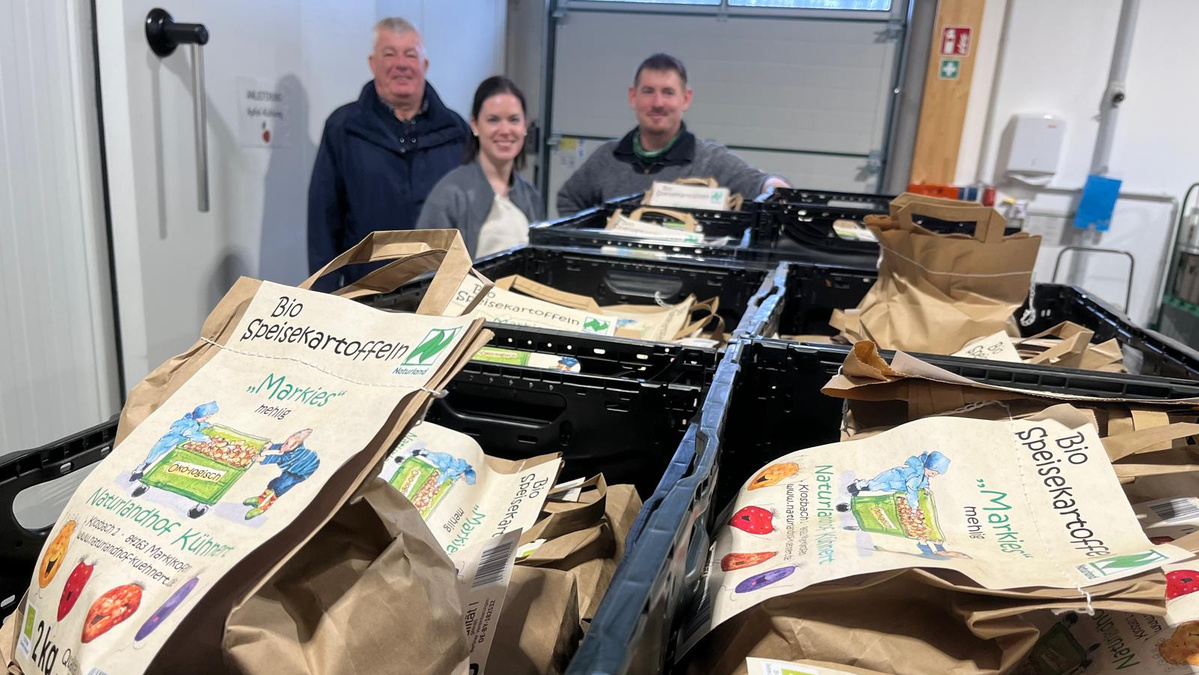 KI generiert: Das Bild zeigt drei Personen, die hinter mehreren Kisten mit braunen Papiertüten voller Bio-Speisekartoffeln stehen. Die Tüten beschreiben verschiedene Sorten und Mengen von Kartoffeln.