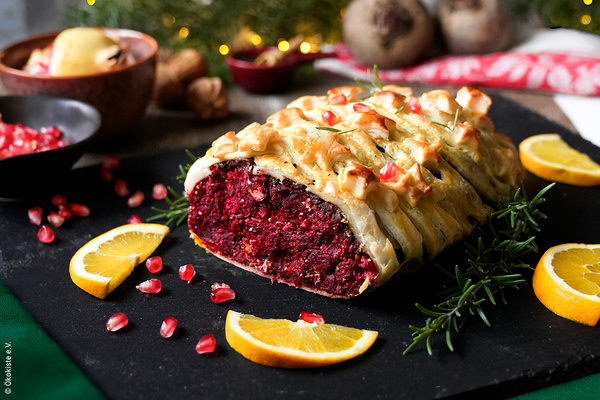 KI generiert: Das Bild zeigt ein veganes Wellington aus Roter Bete, das kunstvoll in Blätterteig eingewickelt und mit Granatapfelkernen sowie Rosmarin dekoriert ist. Es ist von Orangenscheiben umgeben und auf einer Schieferplatte angerichtet.