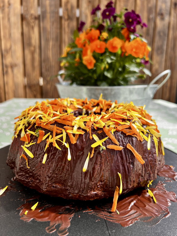 KI generiert: Ein Schokoladenkuchen mit bunter Blüten- und Karottendekoration vor einem Blumengesteck.