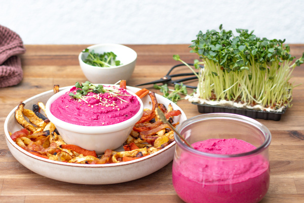 KI generiert: Das Bild zeigt einen veganen Burger mit grünen Zutaten und Brötchen mit Sesam, serviert auf einem Holzuntergrund. Im Hintergrund sind weitere Burgerzutaten, ein kleiner Napf mit Dip und eine Schüssel mit Brötchen zu sehen.