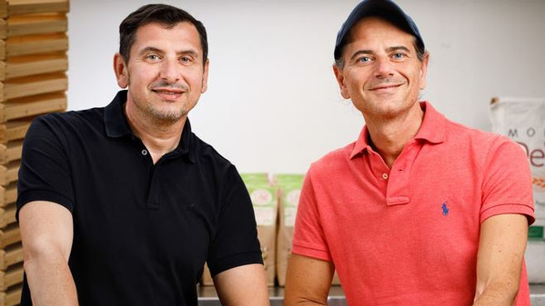 KI generiert: Zwei lächelnde Männer sitzen nebeneinander. Im Hintergrund sind Verpackungen zu sehen.