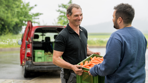 KI generiert: Zwei Männer beim Austausch einer Kiste mit Karotten vor einem Auto im Freien.