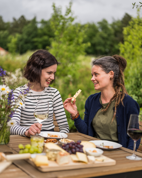 KI generiert: Zwei Personen genießen im Freien Wein und Käse an einem Tisch mit Blumen und einer ländlichen Kulisse.