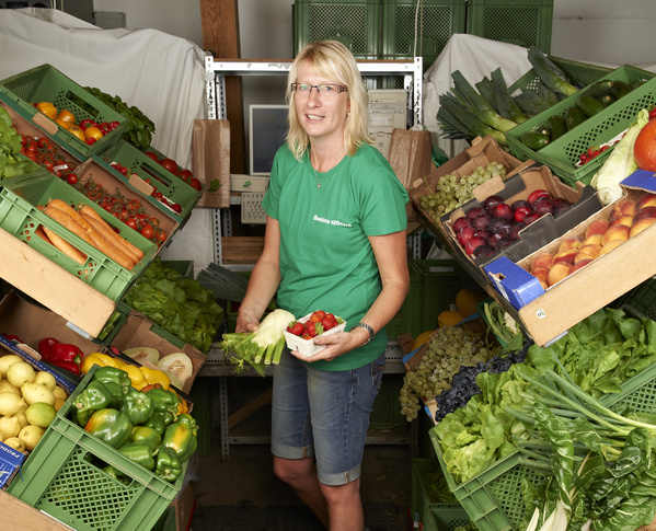 KI generiert: Frau in Obst- und Gemüselager mit Körbchen voller Erdbeeren, umgeben von frischen Produkten.