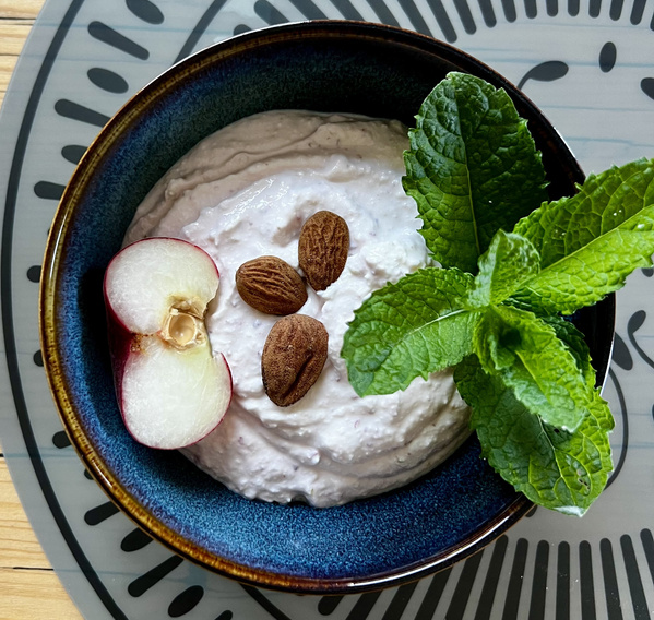 KI generiert: Blaue Schüssel mit Joghurt, Mandeln, halbem Pfirsich und Minzblättern auf gemustertem Untersetzer.