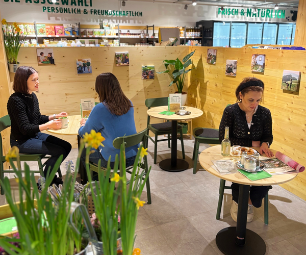 KI generiert: Menschen in einem Café; entspannte Gespräche und Lektüre. Im Hintergrund: "Frisch & natürlich".