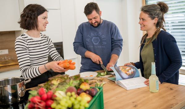 KI generiert: Drei Personen kochen gemeinsam in einer Küche, lesen ein Rezeptbuch und benutzen frisches Gemüse.