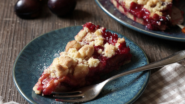 KI generiert: Stück Pflaumenstreuselkuchen auf einem blauen Teller mit Gabel. Im Hintergrund sind einige Pflaumen zu sehen.
