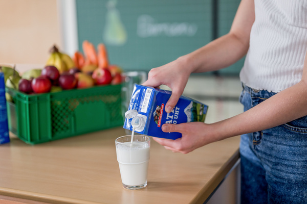 KI generiert: Person füllt ein Glas mit Milch; Hintergrund: Obstkiste. Text auf Tafel: "Ein...".