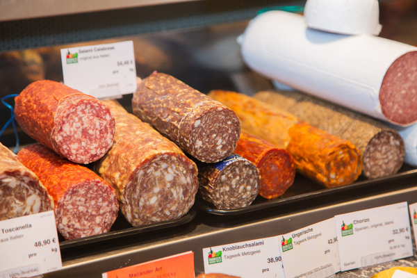 KI generiert: Das Bild zeigt eine Auswahl verschiedener Wurstsorten in einer Metzgerei, darunter Salami und andere Spezialitäten. Sie sind auf einem Display mit entsprechenden Preisschildern präsentiert.