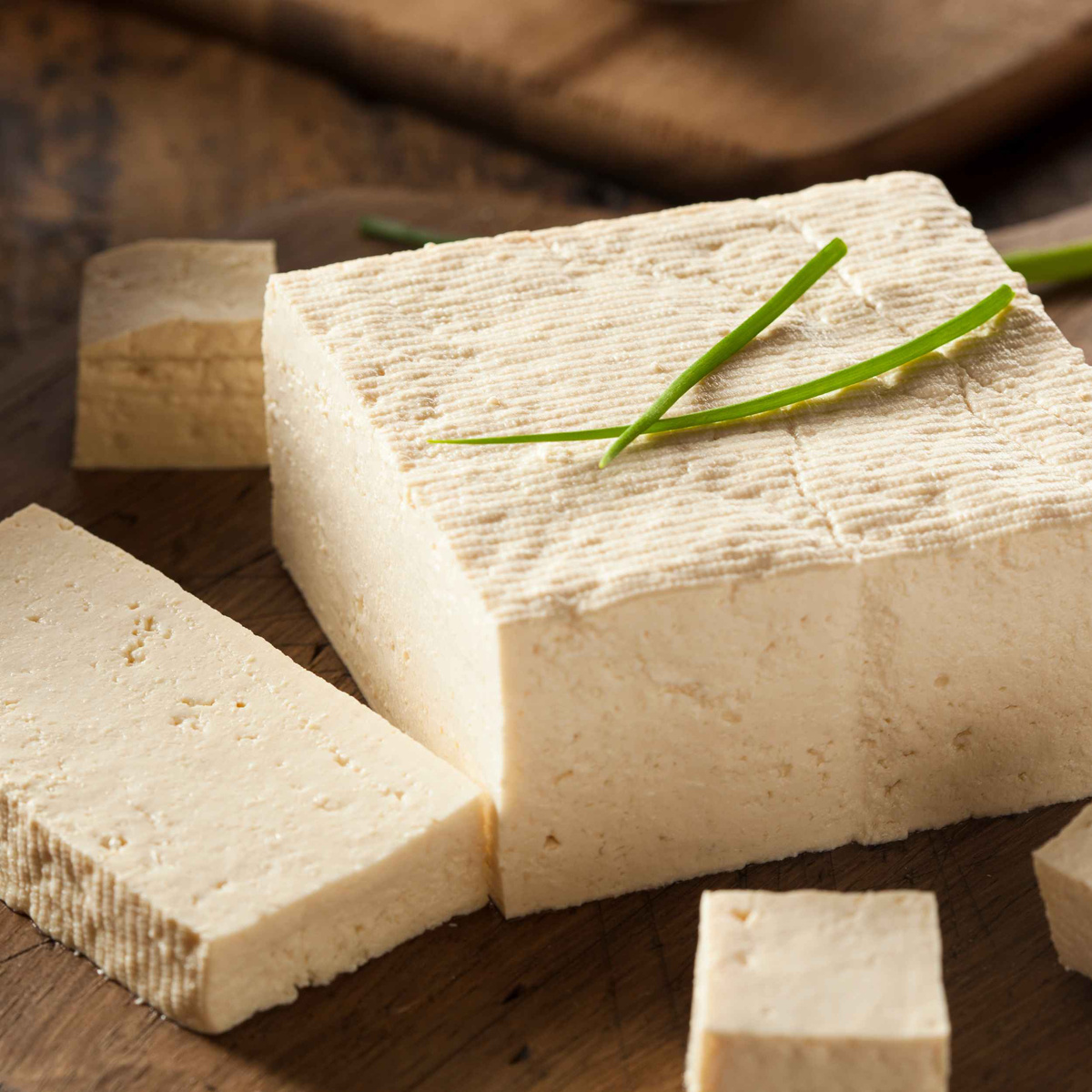 KI generiert: Das Bild zeigt einen Block Tofu, der auf einem Holzbrett liegt, mit einigen geschnittenen Stücken und dekoriert mit Schnittlauch. Der Tofu hat eine cremige Farbe und eine feste Textur.