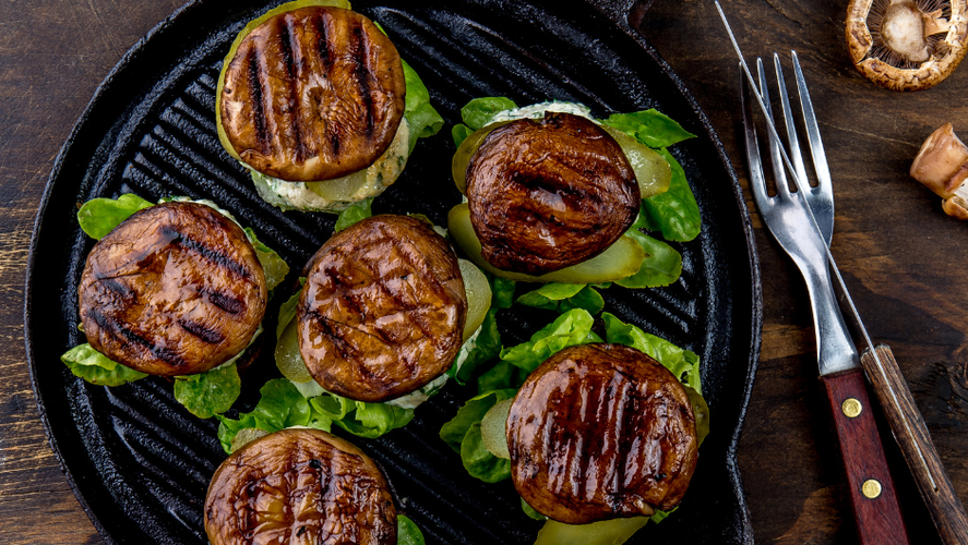 KI generiert: Gegrillte Pilze auf Salatblättern in einer Grillpfanne, daneben liegen Messer und Gabel auf einem Holztisch.