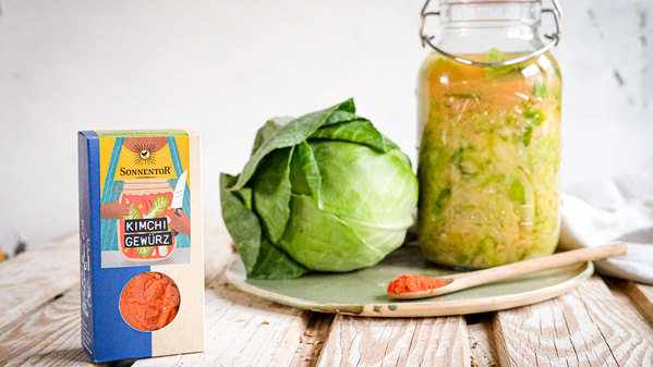 KI generiert: Kimchi-Gewürz, Kohlkopf und Glas mit eingelegtem Kimchi auf einem Tisch. Text: "Sonnentor Kimchi Gewürz".