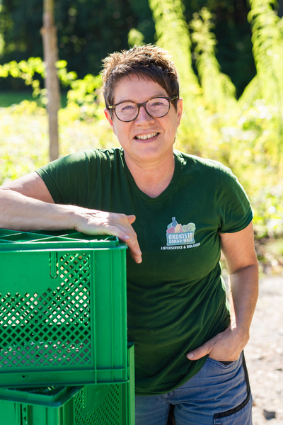 KI generiert: Eine Person lehnt an grünen Kisten und lächelt. Ihr Shirt zeigt das Logo "Ökokiste Donau-Wald".