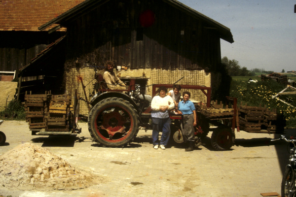 KI generiert: Personen posieren vor einem Traktor mit Anhänger auf einem Bauernhof. Im Hintergrund ist ein Holzgebäude.