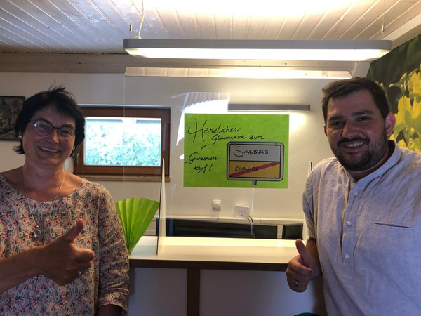 KI generiert: Zwei Personen geben Daumen hoch vor einem Schild: "Herzlichen Glückwunsch zum Grundstückskauf! Saulburg".