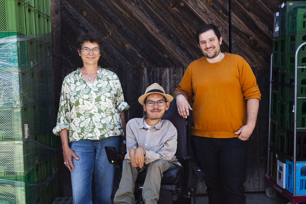 KI generiert: Drei Personen stehen lächelnd vor Holzwand und grünen Kisten.