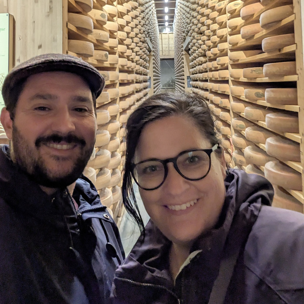 KI generiert: Zwei Personen lächeln in einem Raum voller gelagerter Käselaibe.