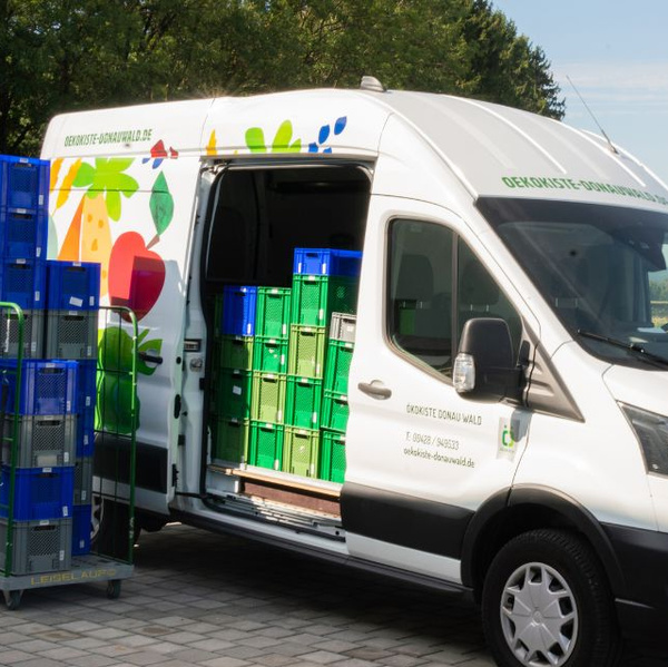 KI generiert: Weißer Lieferwagen mit buntem Gemüse-Design, beladen mit grünen Kisten. Text: "Oekokiste-Donauwald.de".