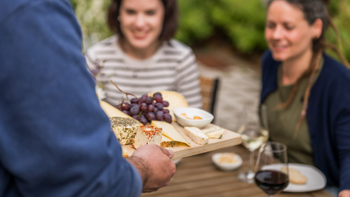 KI generiert: Person serviert Käseplatte mit Weintrauben und Dips; zwei Frauen lächeln am Tisch.
