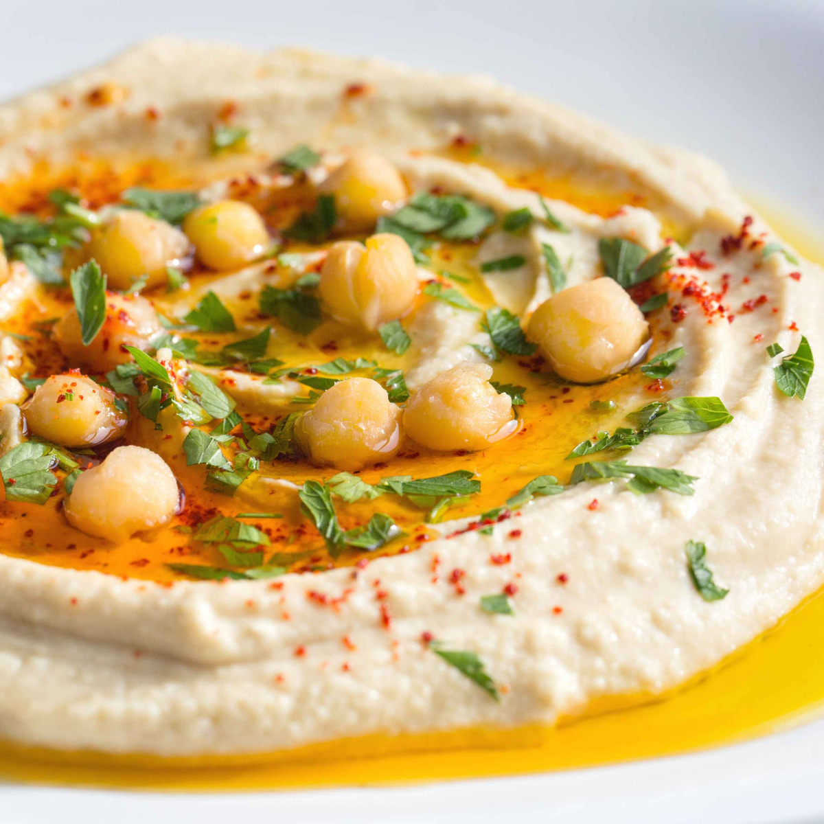 KI generiert: Das Bild zeigt eine Portion Hummus, garniert mit Kichererbsen, Olivenöl, gehackter Petersilie und Paprikapulver. Das Gericht ist auf einem Teller angerichtet.