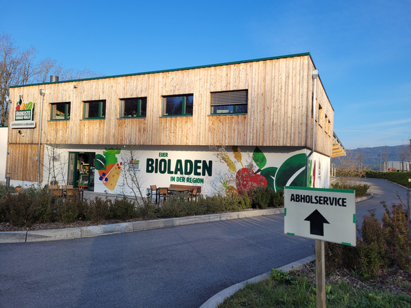KI generiert: Holzgebäude eines Bioladens mit "ABHOLSERVICE"-Schild. Texto: "Euer Bioladen in der Region".