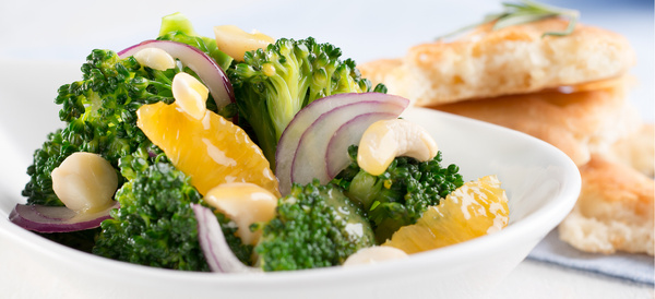 KI generiert: Das Bild zeigt einen frischen Brokkoli-Salat mit Orangenscheiben, roten Zwiebeln und Cashewnüssen in einer weißen Schale. Im Hintergrund sind warme Brotscheiben zu sehen.