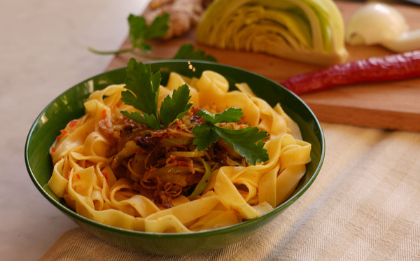 KI generiert: Ein Teller mit Nudeln und Gemüse, im Hintergrund Zutaten wie Lauch und Paprika auf einem Schneidebrett.